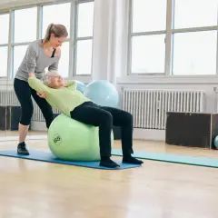Une femme âgée fait des exercices pour renforcer son muscle du dos sur un ballon de gym, avec l'aide d'une coach.