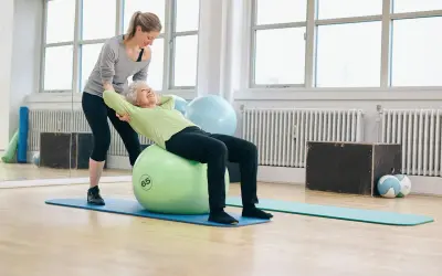 Une femme âgée fait des exercices pour renforcer son muscle du dos sur un ballon de gym, avec l'aide d'une coach.