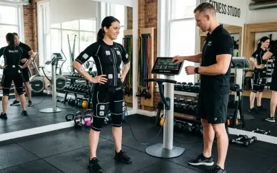 Femme en combinaison EMS, prête pour un exercice pour perdre du ventre, guidée par un coach devant une machine.