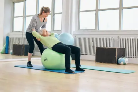 Une femme âgée fait des exercices pour renforcer son muscle du dos sur un ballon de gym, avec l'aide d'une coach.