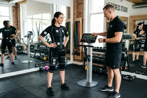 Femme en combinaison EMS, prête pour un exercice pour perdre du ventre, guidée par un coach devant une machine.