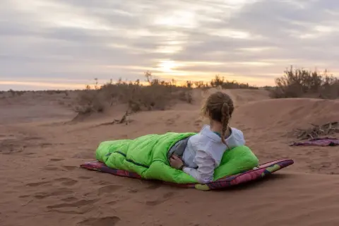 Fin du **trek dans le désert**. Une jeune femme en sac de couchage vert admire le coucher de soleil sur les dunes.