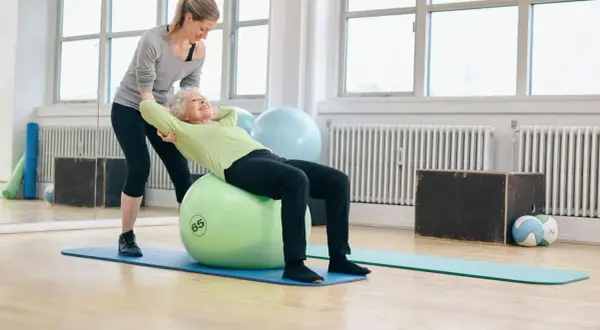 Une femme âgée fait des exercices pour renforcer son muscle du dos sur un ballon de gym, avec l'aide d'une coach.