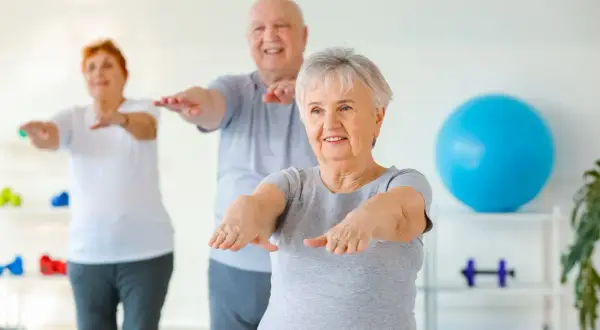 Trois seniors souriants pratiquent la gym chinoise pour seniors, bras tendus, dans une salle lumineuse.