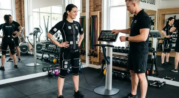 Femme en combinaison EMS, prête pour un exercice pour perdre du ventre, guidée par un coach devant une machine.
