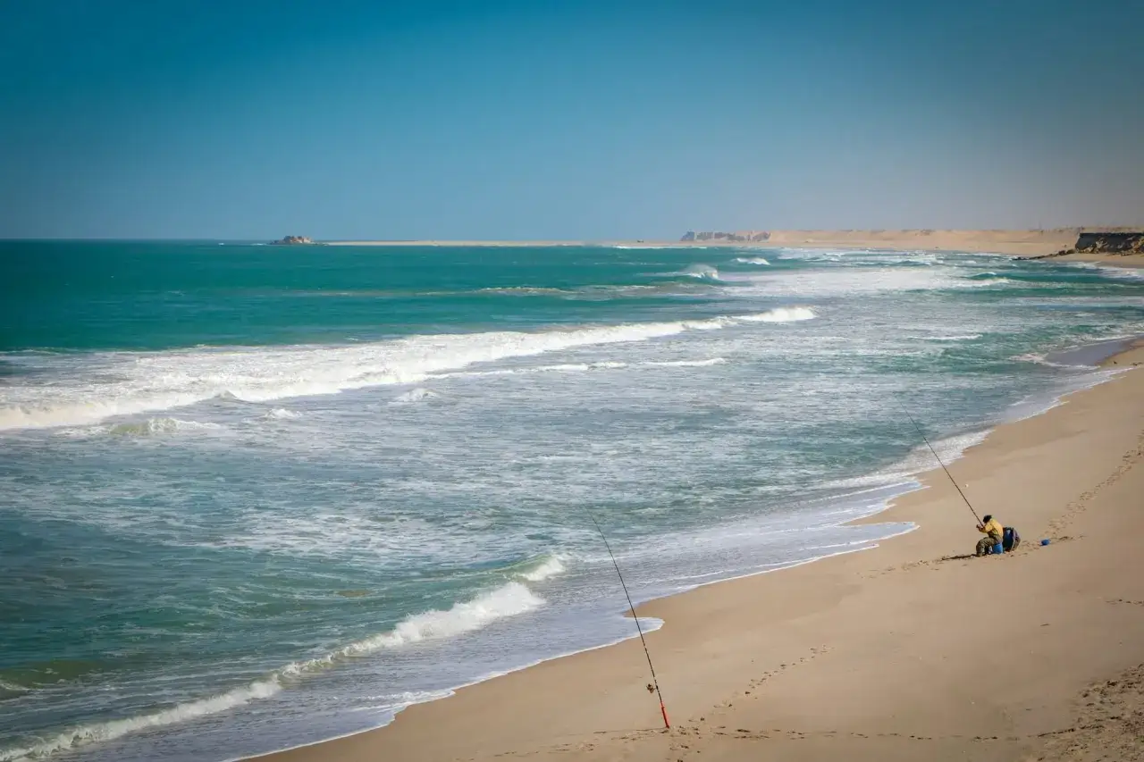 Pêcheur solitaire sur la plage de Dakhla, Maroc, profitant des vagues turquoise sous un ciel bleu.