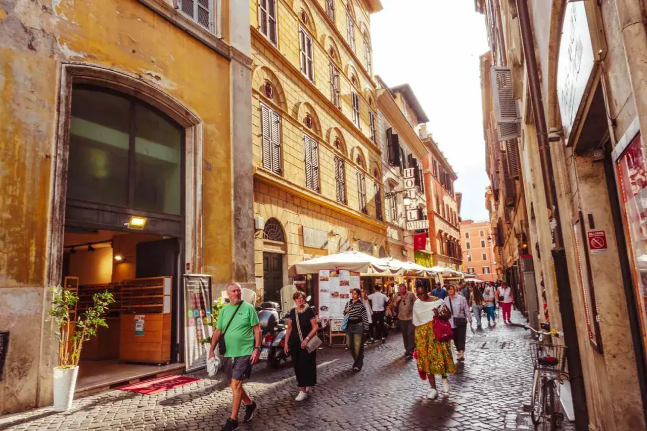 Une rue pav&eacute;e anim&eacute;e &agrave; Rome, une destination europ&eacute;enne de r&ecirc;ve. Des b&acirc;timents anciens, des terrasses de caf&eacute; et des passants cr&eacute;ent une atmosph&egrave;re vibrante.