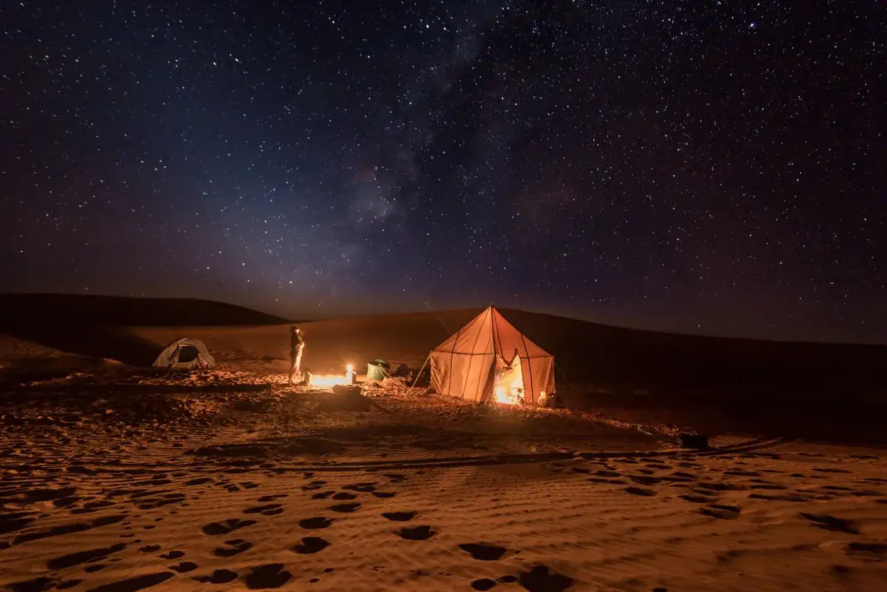 Une nuit dans le d&eacute;sert marocain, sous un ciel &eacute;toil&eacute;. Un campement avec une tente illumin&eacute;e et un feu de camp r&eacute;chauffe le sable.