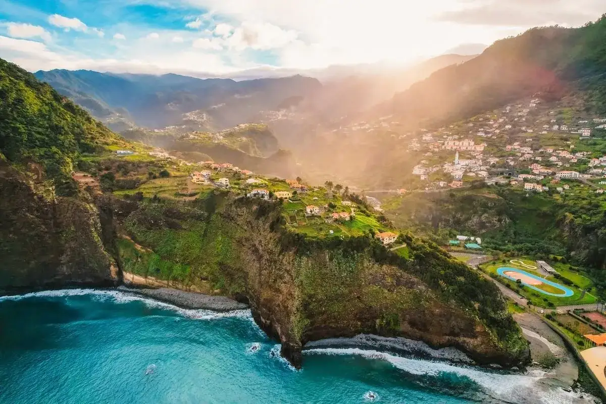 Vue aérienne de falaises verdoyantes plongeant dans l'océan bleu, avec un village pittoresque accroché à flanc de colline. Un décor idyllique pour savoir quand partir à Madère.