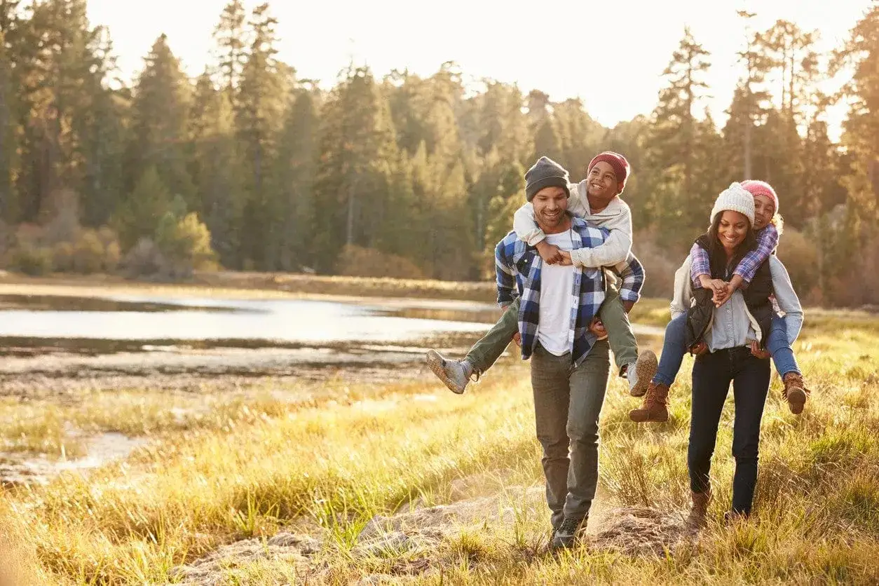 Un couple porte ses enfants sur son dos, profitant d'une micro aventure en famille dans un paysage naturel baign&eacute; de soleil.