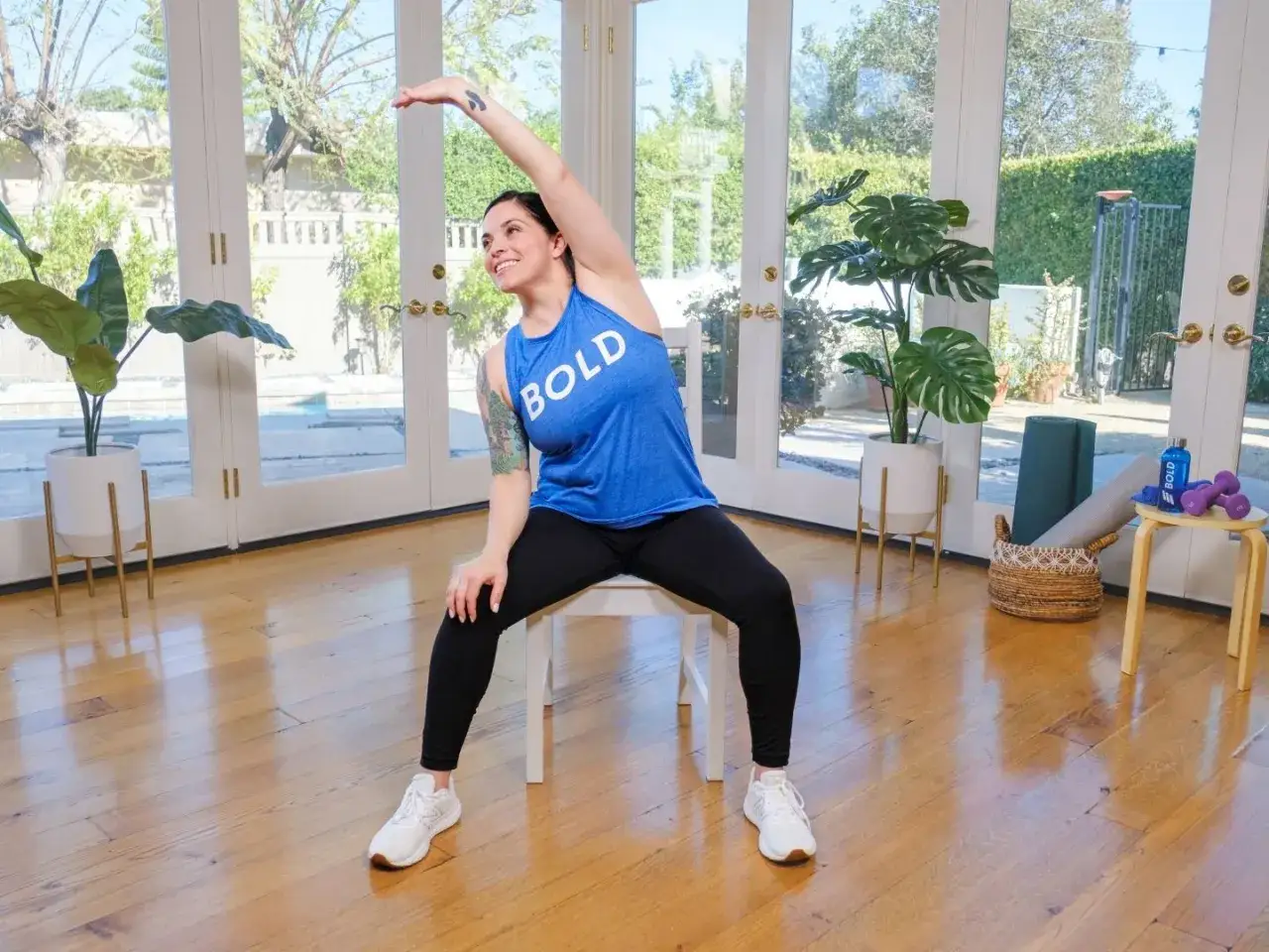 Femme souriante faisant des exercices de pilates gratuit pour seniors, assise sur une chaise, bras levé.