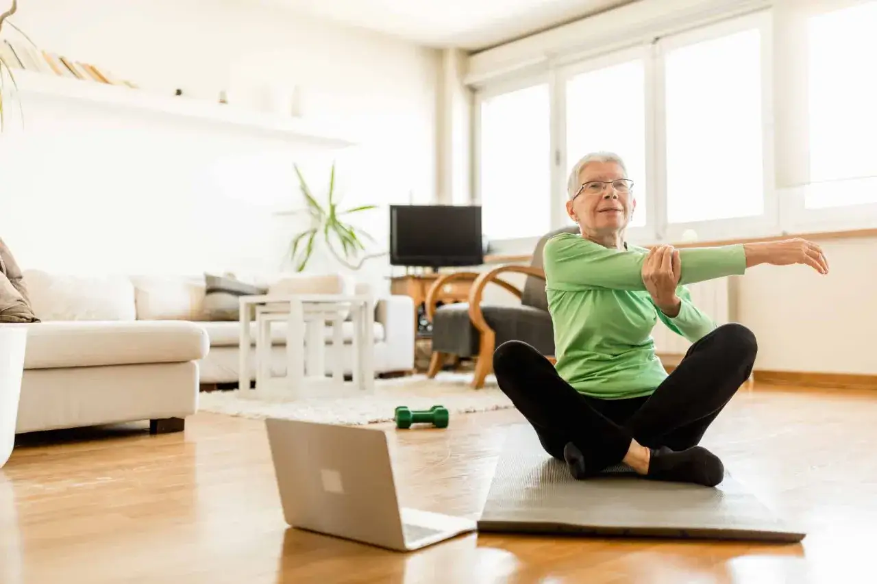 Une femme âgée s'étire chez elle, un ordinateur portable ouvert devant elle, prouvant qu'il est possible de prendre du muscle après 50 ans.
