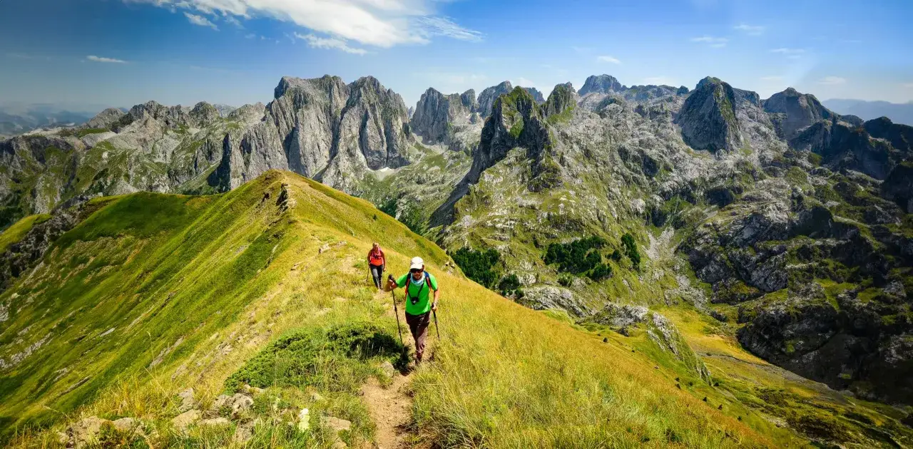 Deux randonneurs profitent d'un trek en Albanie, traversant une crête herbeuse avec des montagnes majestueuses en arrière-plan.