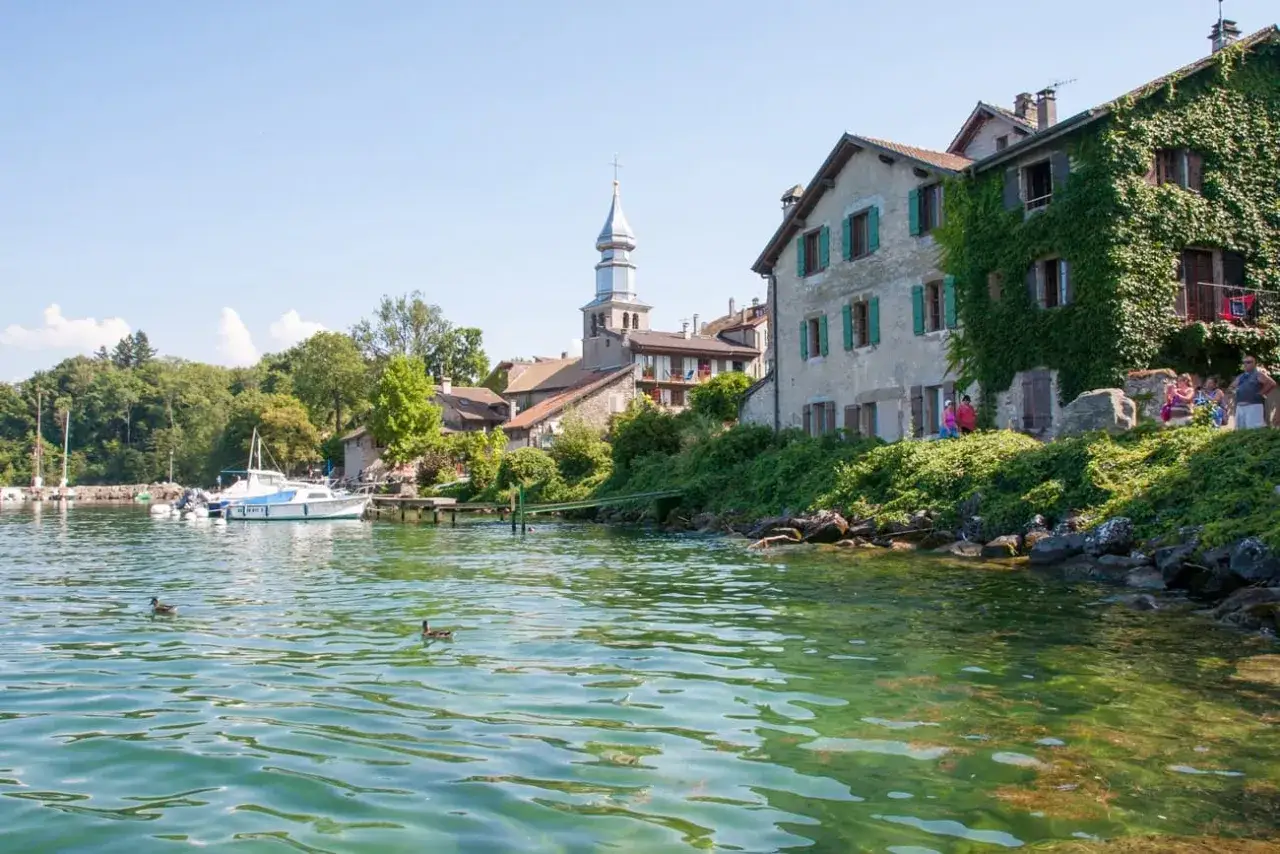 Bateaux amarrés, maisons fleuries et clocher se reflétant dans l'eau turquoise. Parfaites vacances Alpes été.