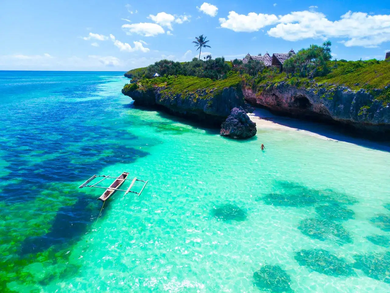 Un paradis tropical en Tanzanie, id&eacute;al pour savoir quand partir en Tanzanie. Eau turquoise, pirogue, et une personne nageant pr&egrave;s d'une plage isol&eacute;e.