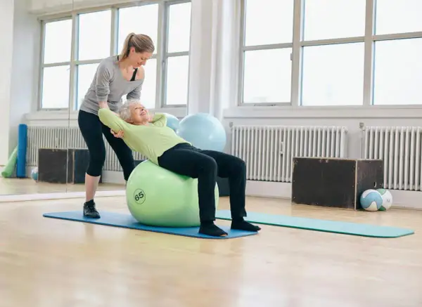 Une femme âgée fait des exercices pour renforcer son muscle du dos sur un ballon de gym, avec l'aide d'une coach.