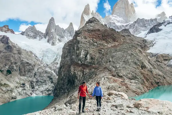 Deux randonneurs se tiennent la main sur un sentier rocheux, admirant un lac turquoise et des montagnes enneigées lors d'une randonnée argentine.