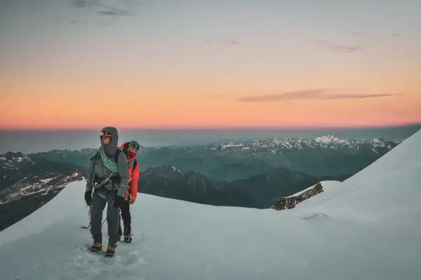 Deux alpinistes entament leur ascension du Mont Rose au lever du soleil, le ciel aux teintes roses et orangées embrassant les sommets enneigés.