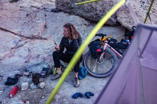 Pause sur une sortie gravel : une cycliste se repose près de son vélo rose, une tasse à la main, au milieu d'un paysage rocheux.